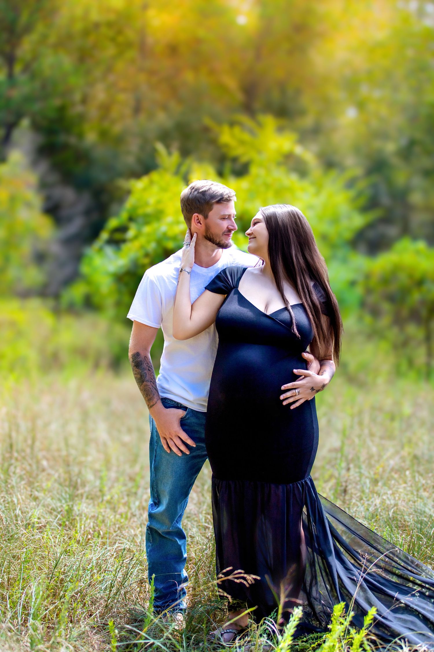 A pregnant couple shares a tender moment outdoors, woman in black gown cradling her belly, man in white shirt embracing her.