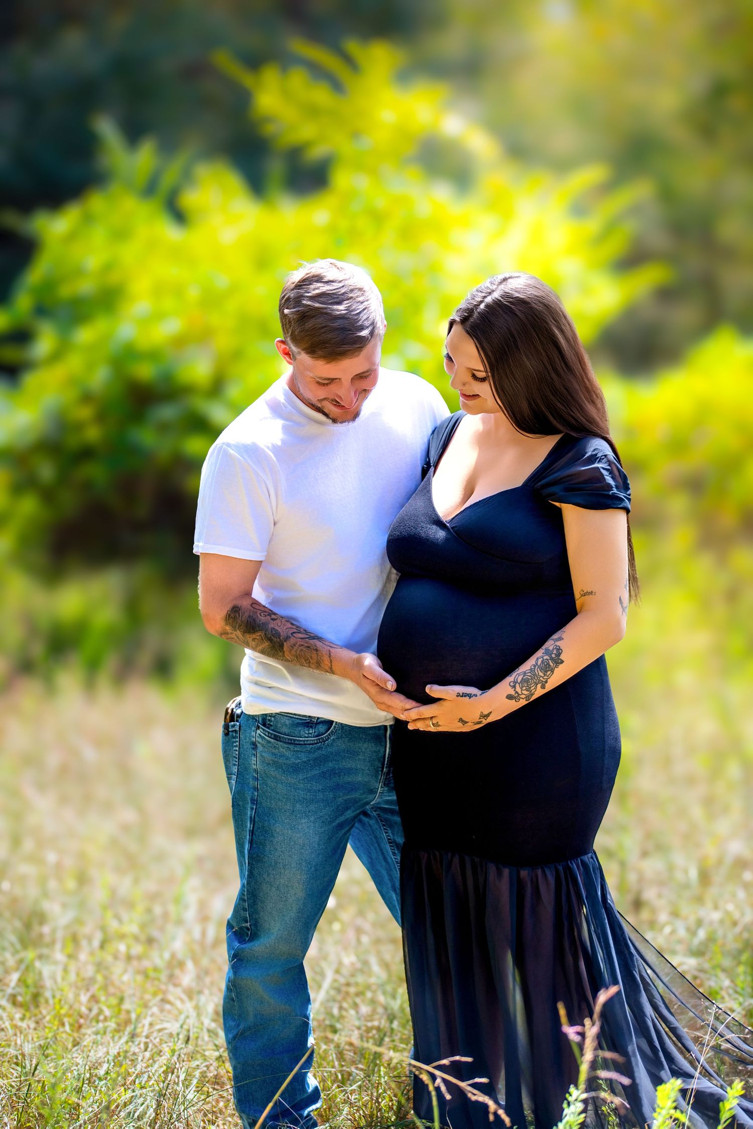 A couple tenderly cradles a pregnant belly outdoors in a sunny field, man in white tee and woman in navy maternity gown.
