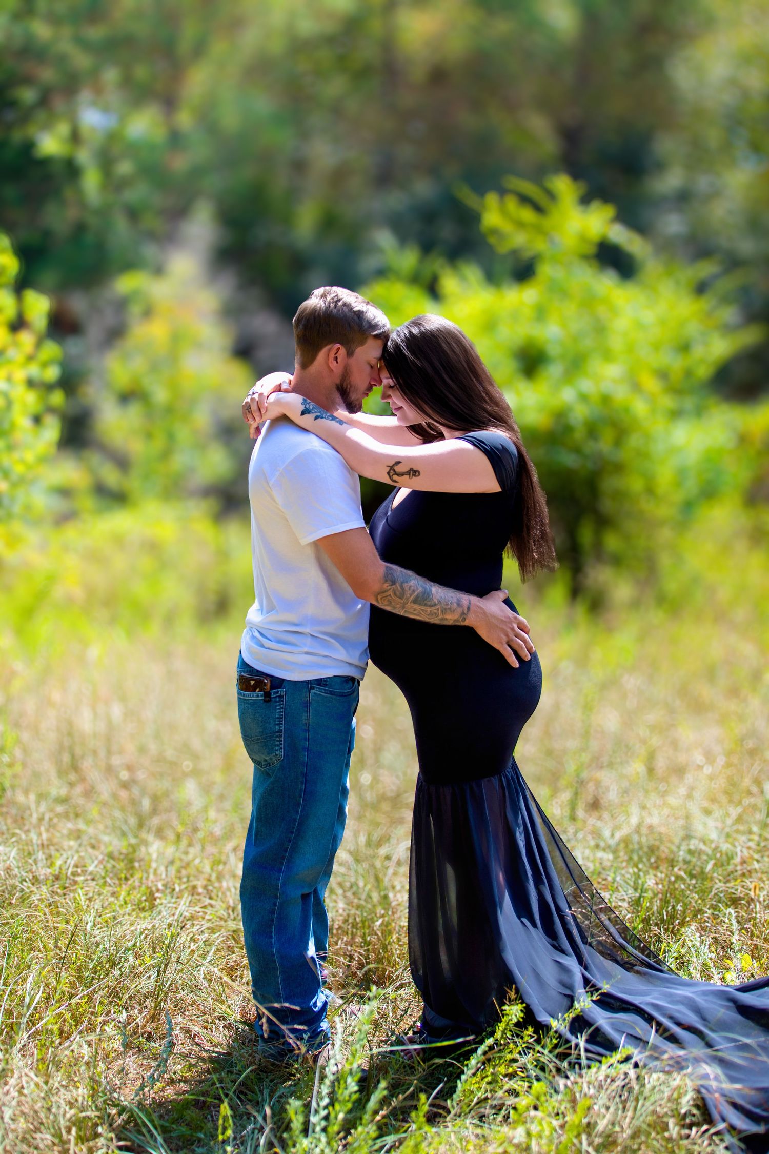 A couple shares a tender moment outdoors, with the pregnant woman in a black gown embracing her partner in a lush green field.