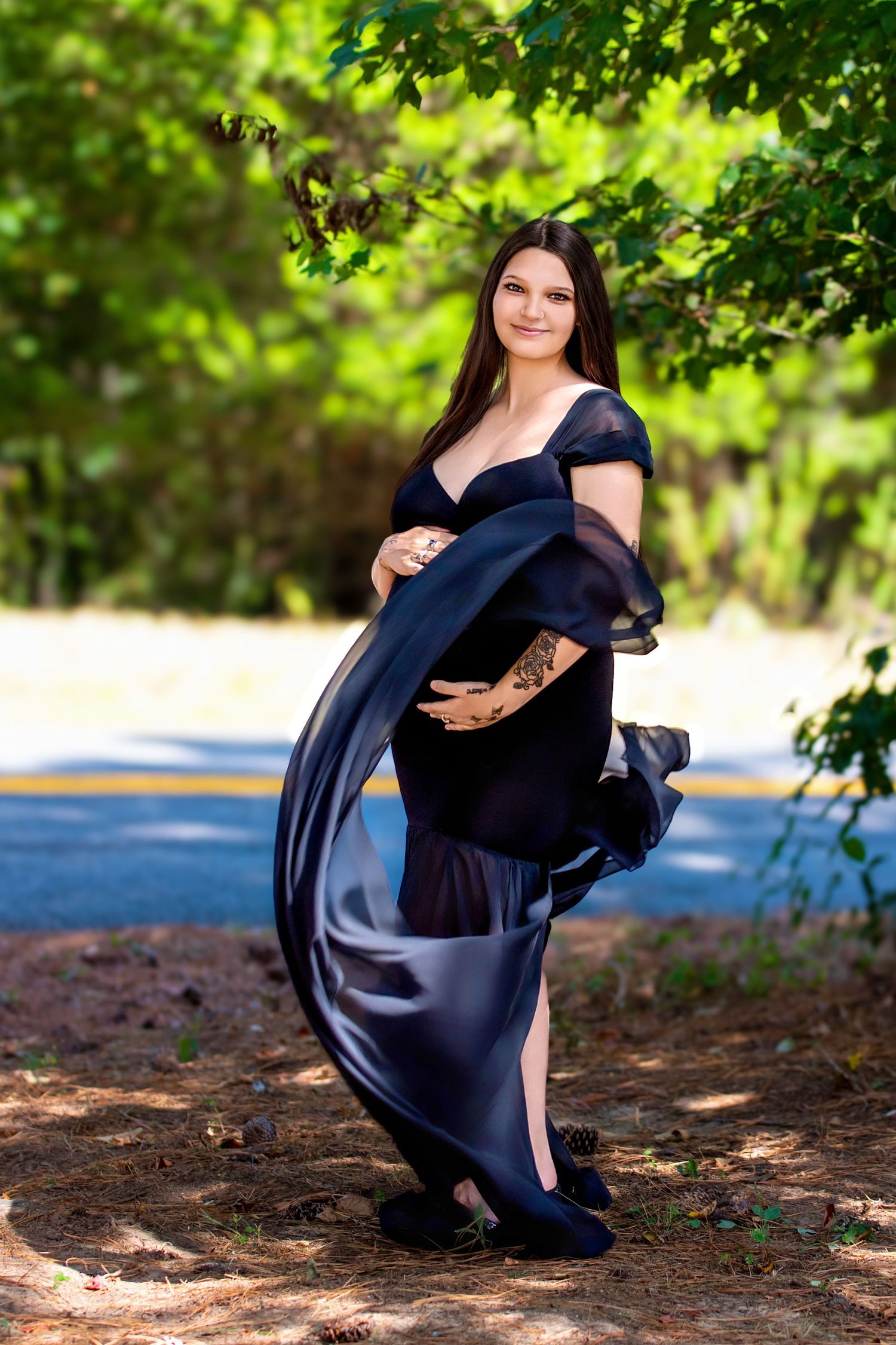 Pregnant woman in flowing navy blue gown posing outdoors among lush green trees during a maternity photo shoot.