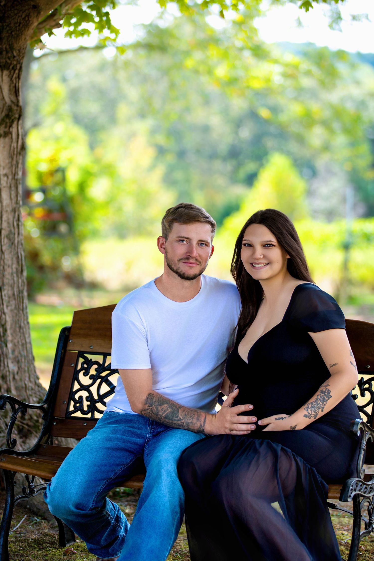 Couple sitting on a wooden bench outdoors, woman in black dress with baby bump, man in white tee and jeans, green trees behind.