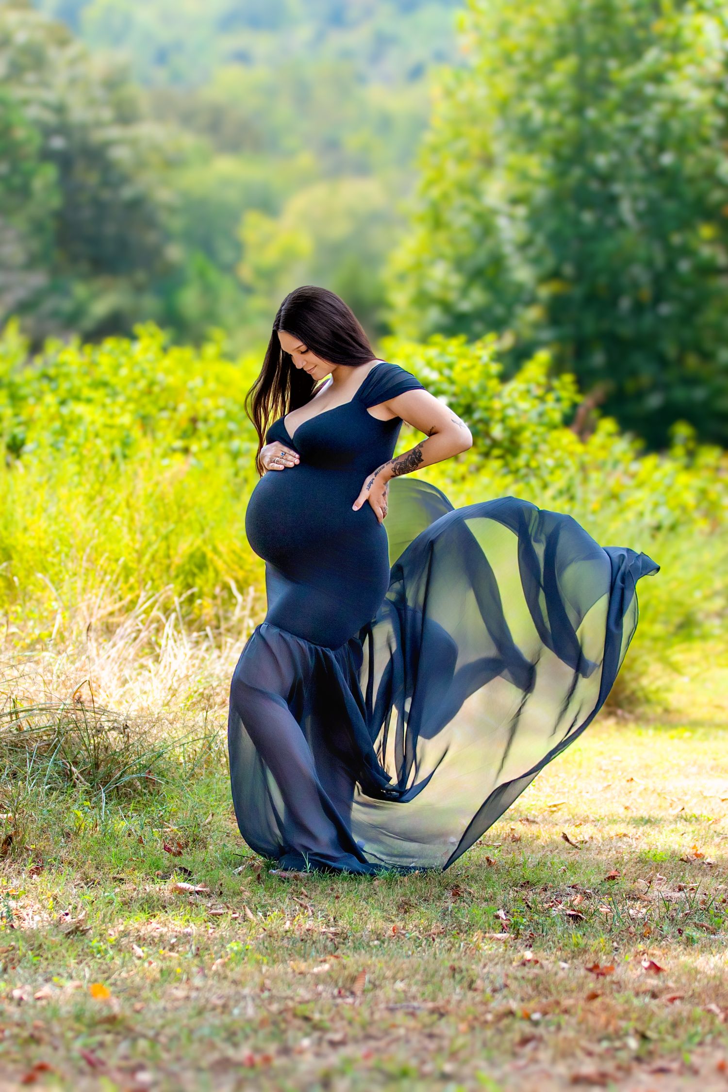 Pregnant woman in flowing navy blue dress standing outdoors in lush green park, cradling baby bump.
