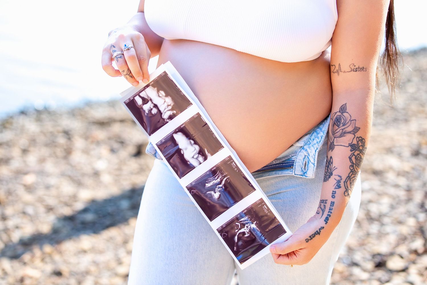 Pregnant woman holding ultrasound photos strip near beach, showing tattooed arm and baby bump in white top and jeans.