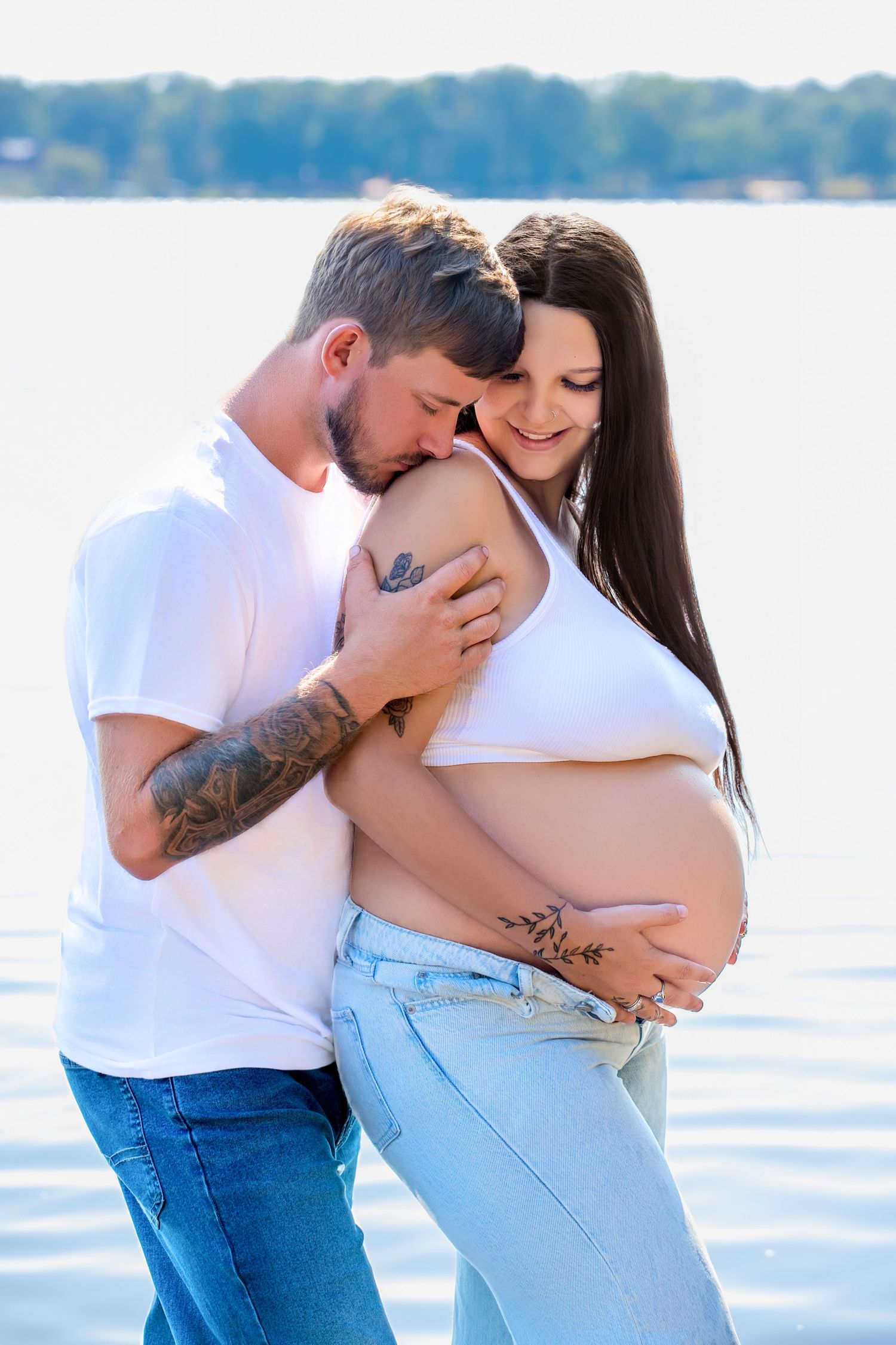 Couple embracing by a lake, pregnant woman in white crop top and jeans, man in white t-shirt with tattoos.