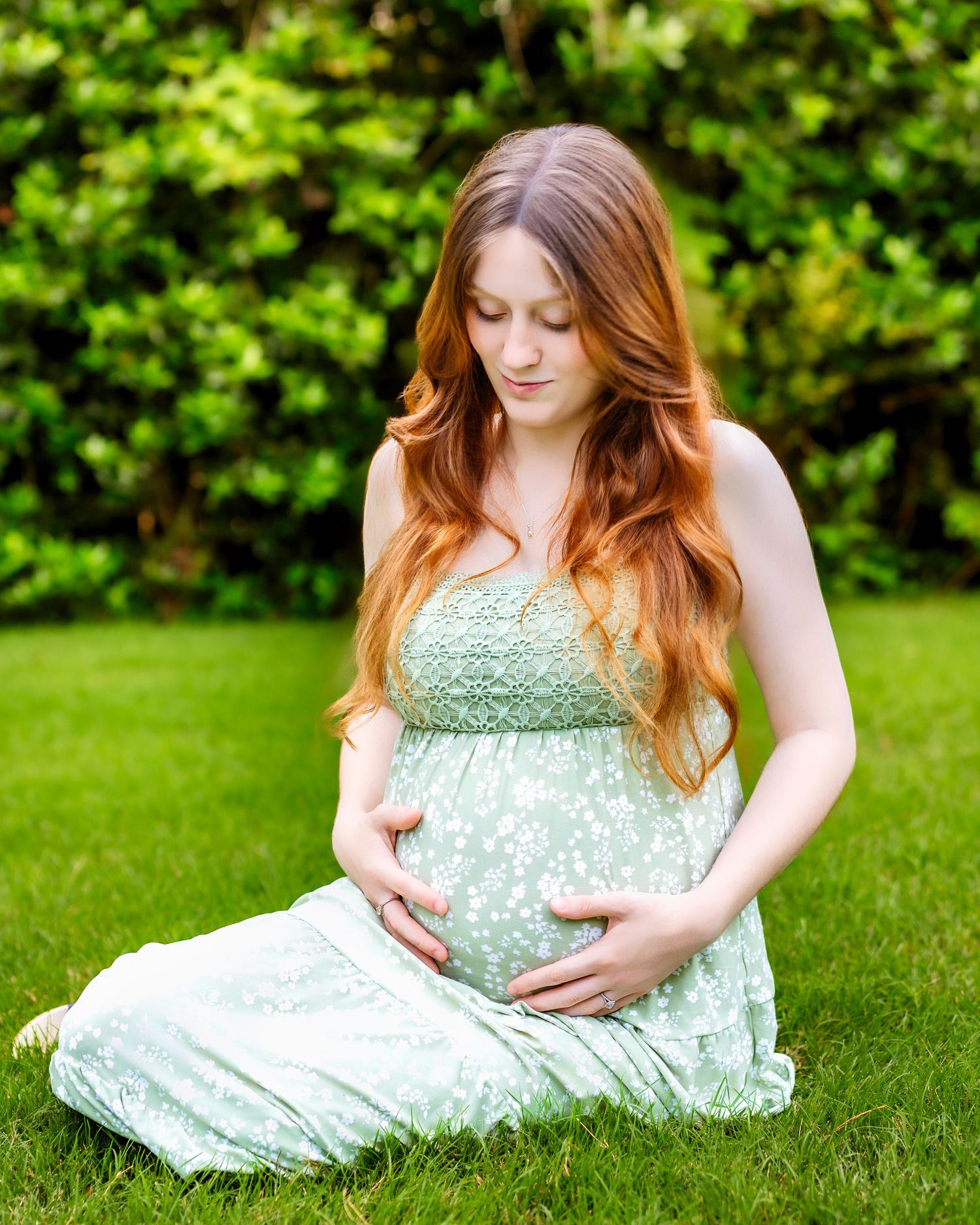 Pregnant woman with long red hair sits on grass in a sage green floral maternity dress, cradling her baby bump.