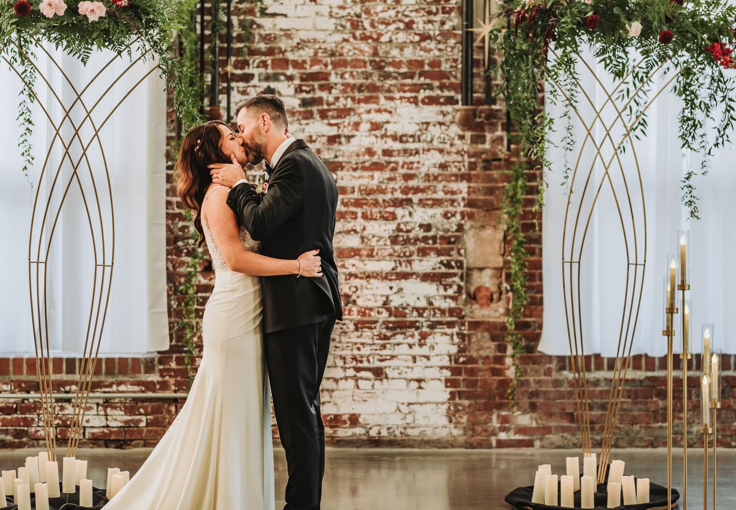 A couple embraces during their wedding ceremony against a rustic exposed brick wall with white arched panels and greenery.
