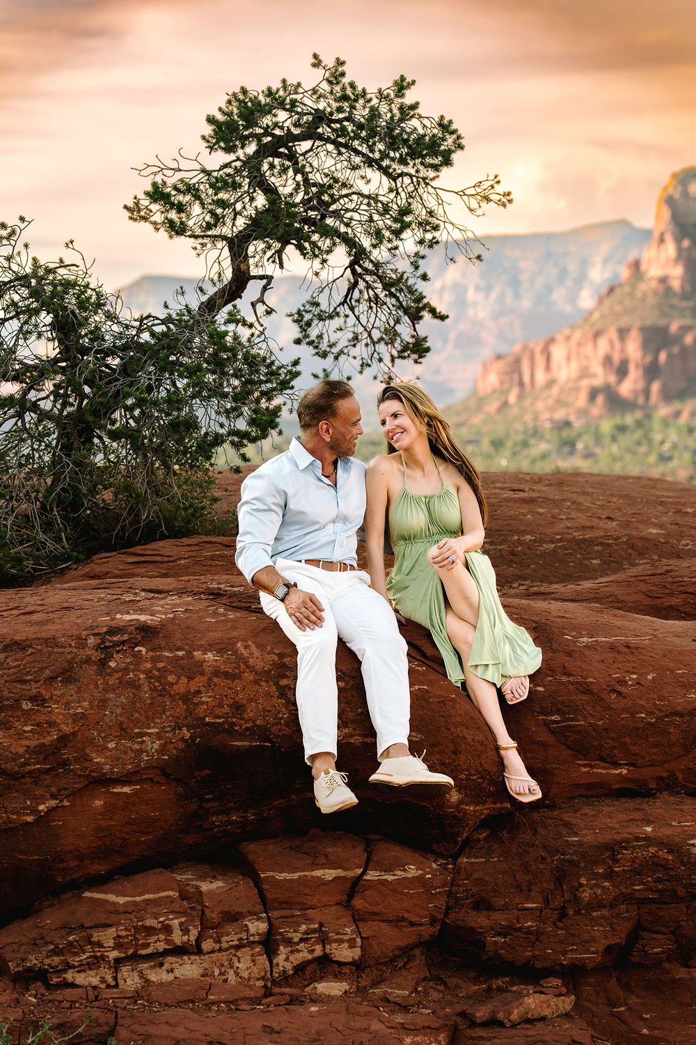 Couple sitting on red rocks in Sedona Arizona at sunset with mountain views and a dramatic pink sky.