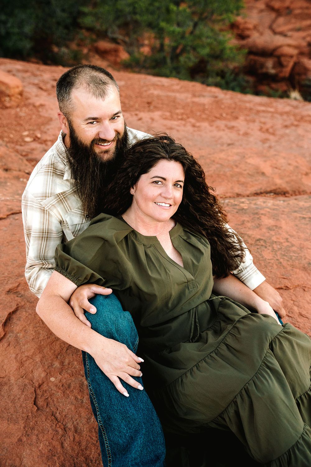 A bearded man and curly-haired woman in a green dress smile together on red rock formations outdoors.