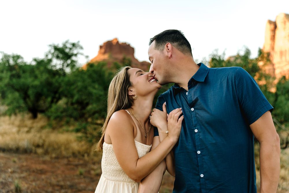 Couple sharing a romantic kiss outdoors in Sedona, Arizona, with red rock formations and green trees in the background.