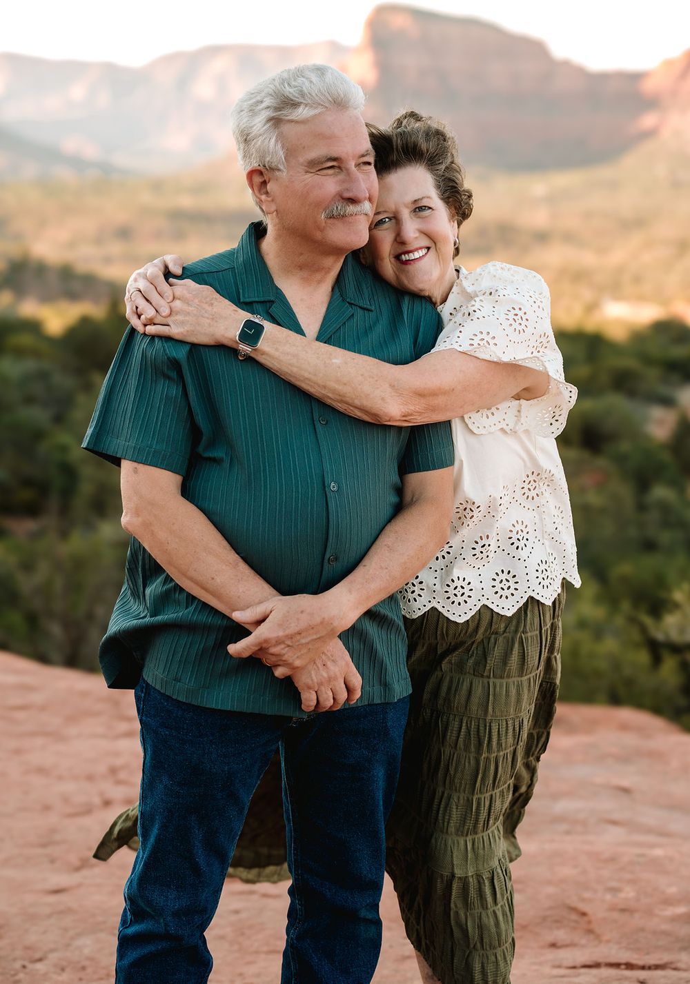Happy older couple hugging on red rock with Sedona mountain backdrop in warm sunset light.