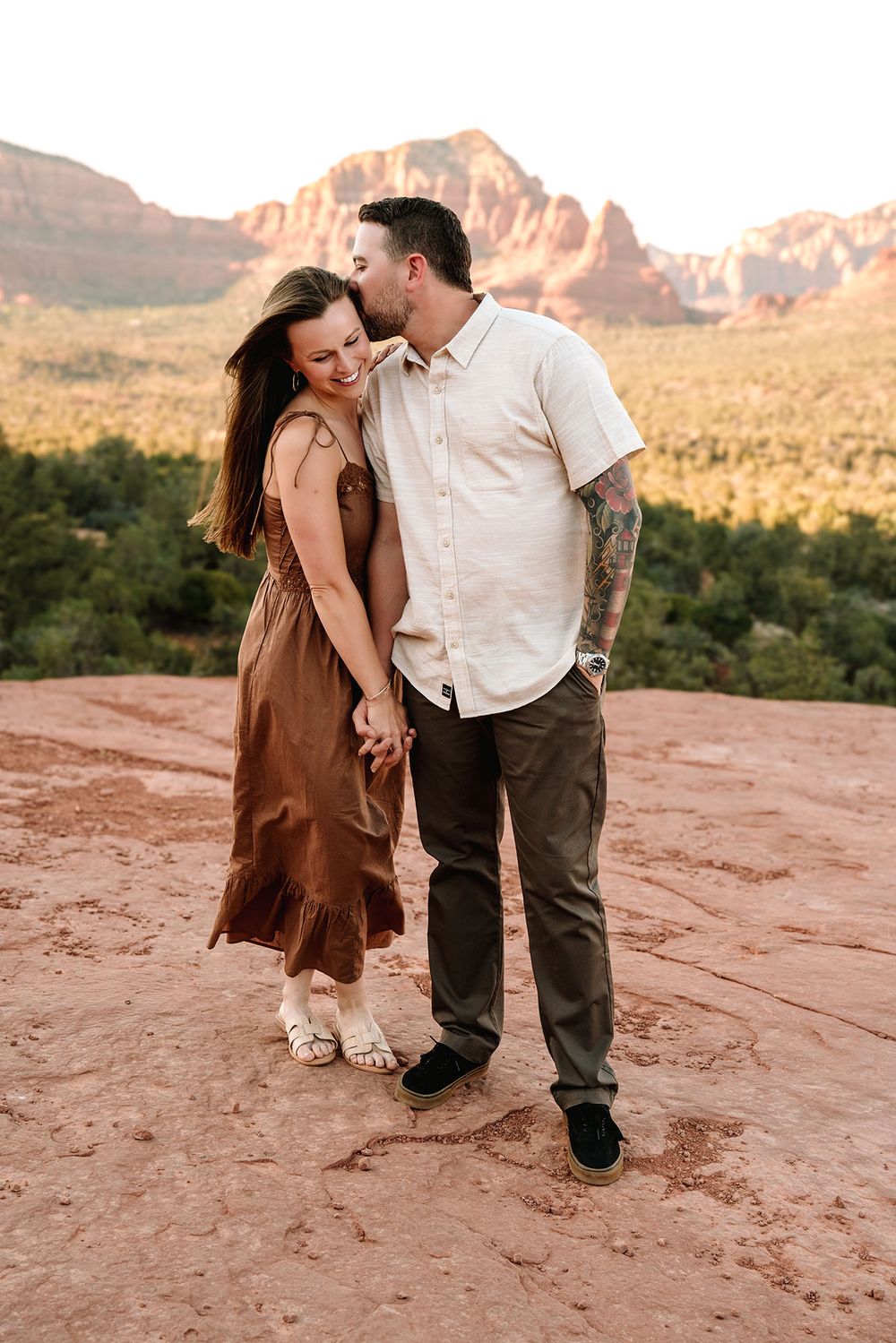 Couple embracing on red rock in Sedona, Arizona, with stunning desert landscape and red mountains at sunset.