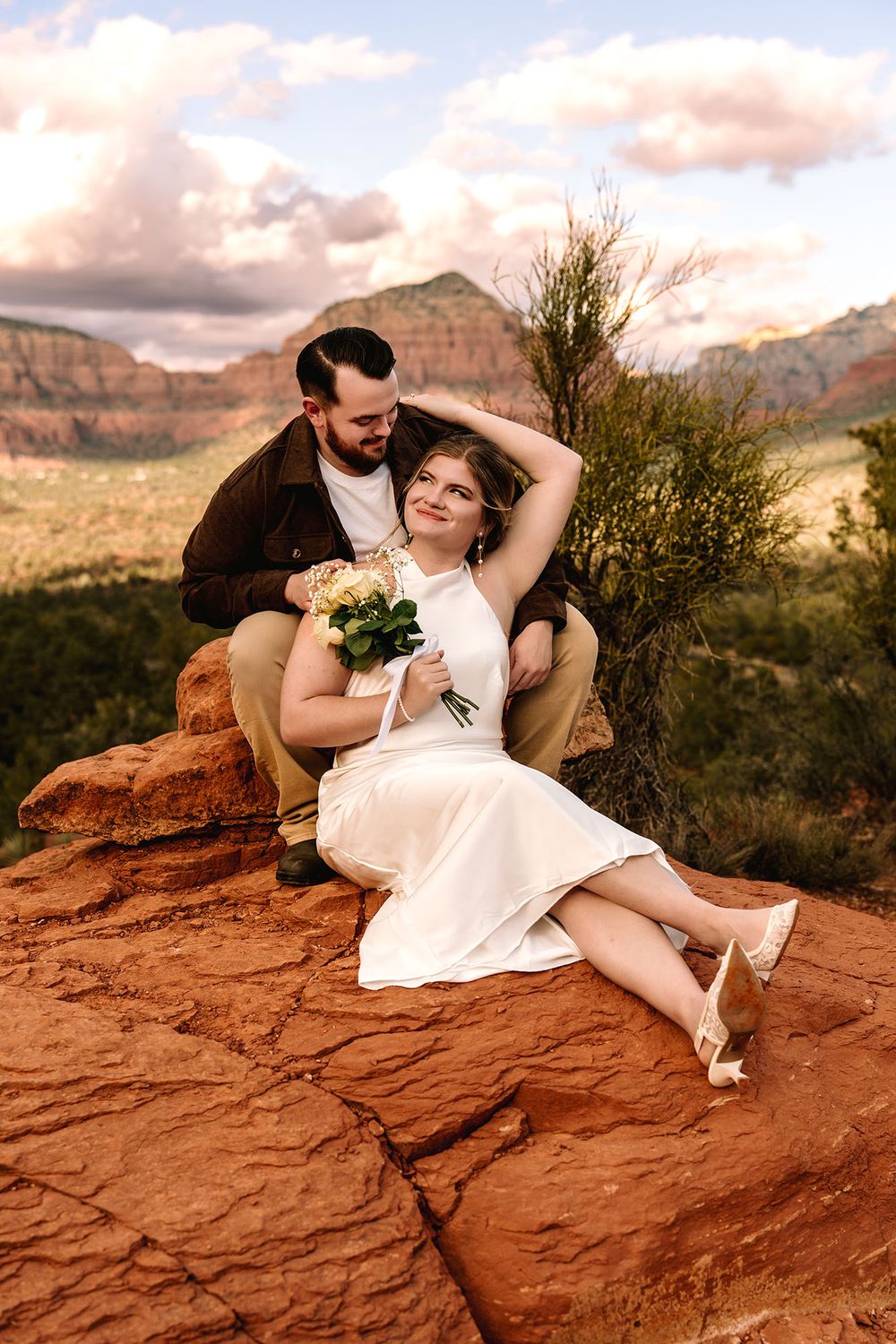 Bride and groom sitting on red rocks in Sedona Arizona with mountain views during their outdoor wedding photo session.