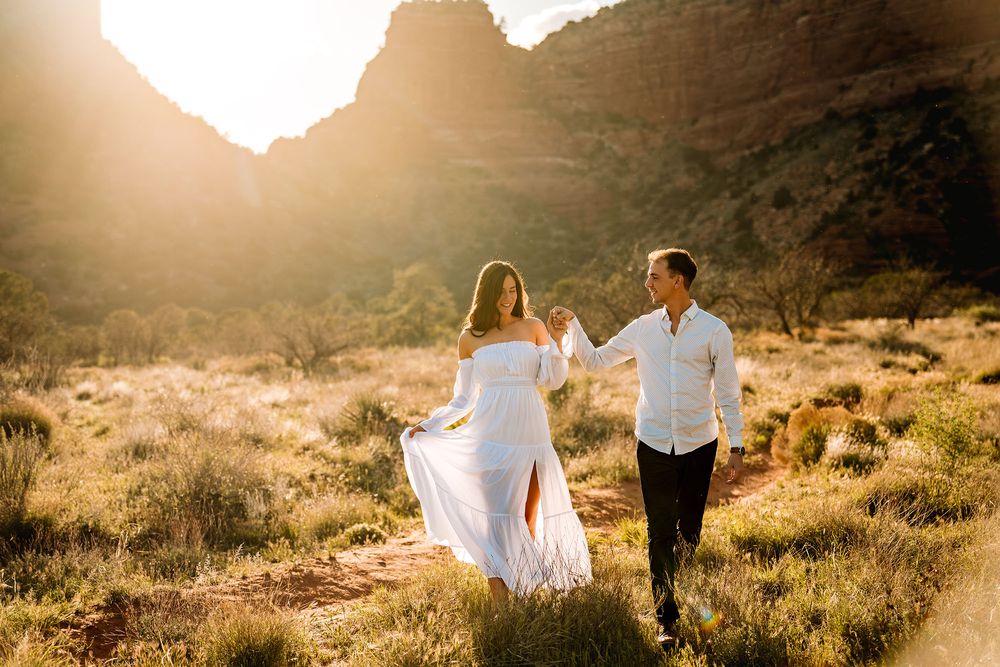 Couple walks hand-in-hand through golden Sedona desert landscape, woman in flowing white dress, mountains glowing at sunset.