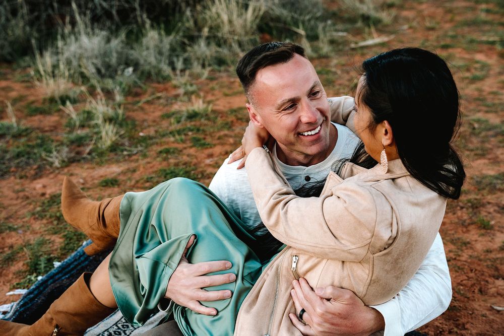 A smiling couple embraces romantically on red desert ground, the man laughing as the woman holds him close outdoors.