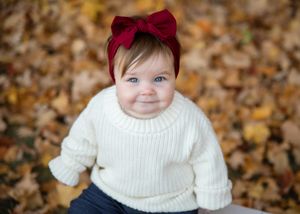 Baby wearing white sweater and red bow on autumn leaves background.