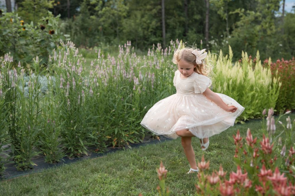 A small figure in a white dress twirls joyfully in a lush garden setting with tall flowers and grass.