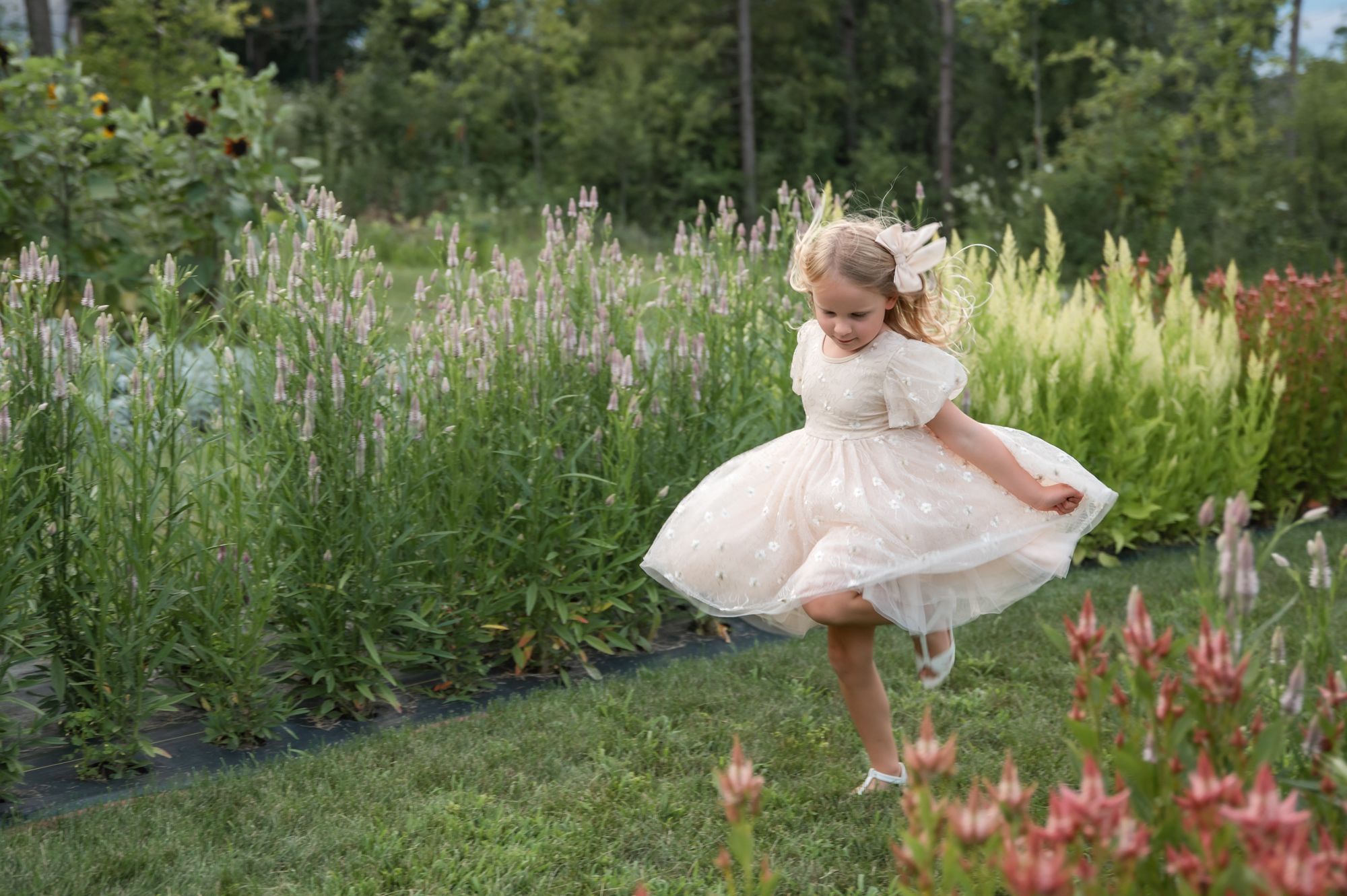 A small figure in a white dress twirls joyfully in a lush garden setting with tall flowers and grass.