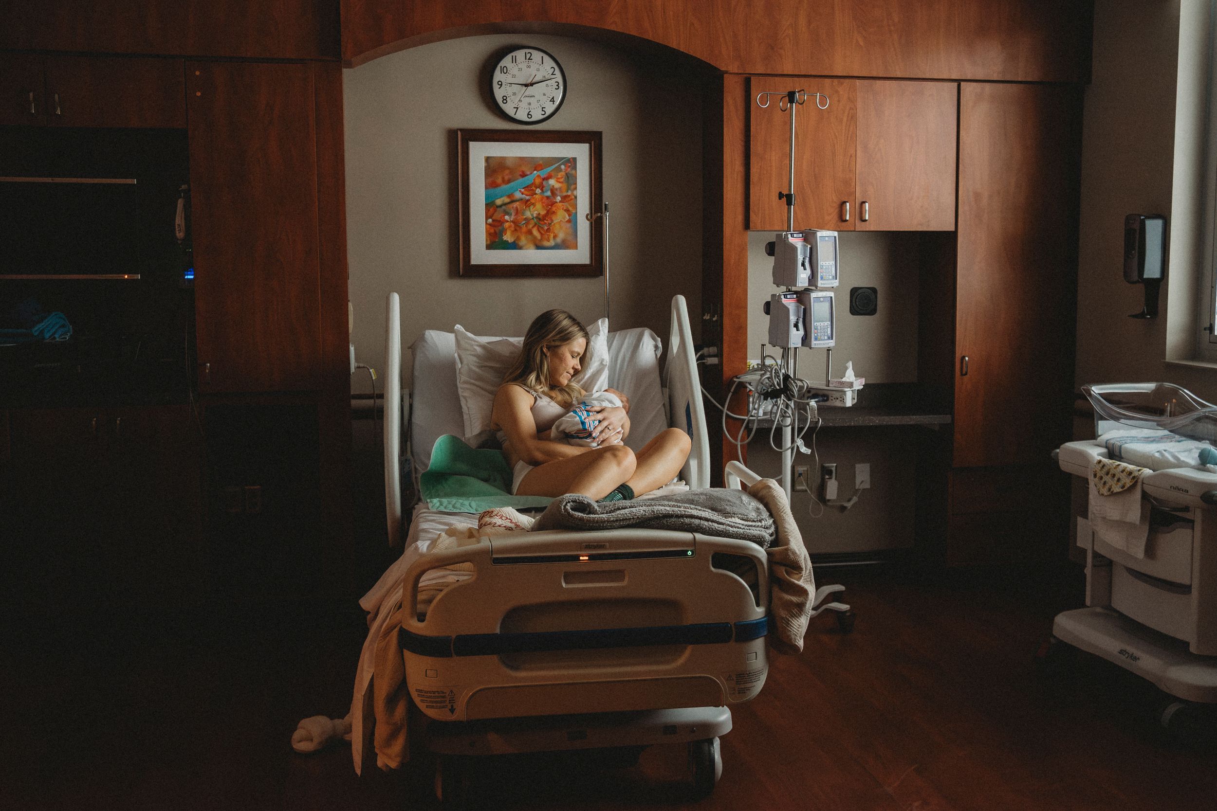 Mother cradles newborn in dimly lit hospital room with wood cabinets and medical equipment.