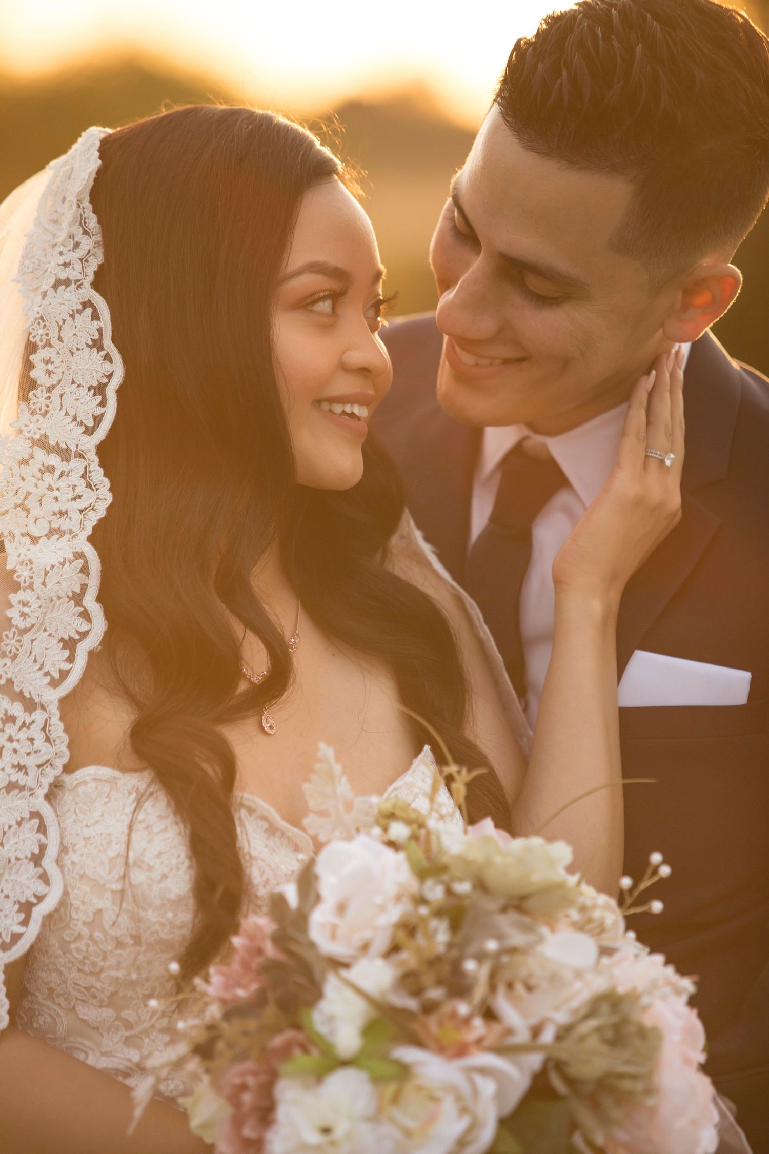 A bride and groom share a romantic golden hour moment, she holds a floral bouquet wearing a lace veil and wedding gown.