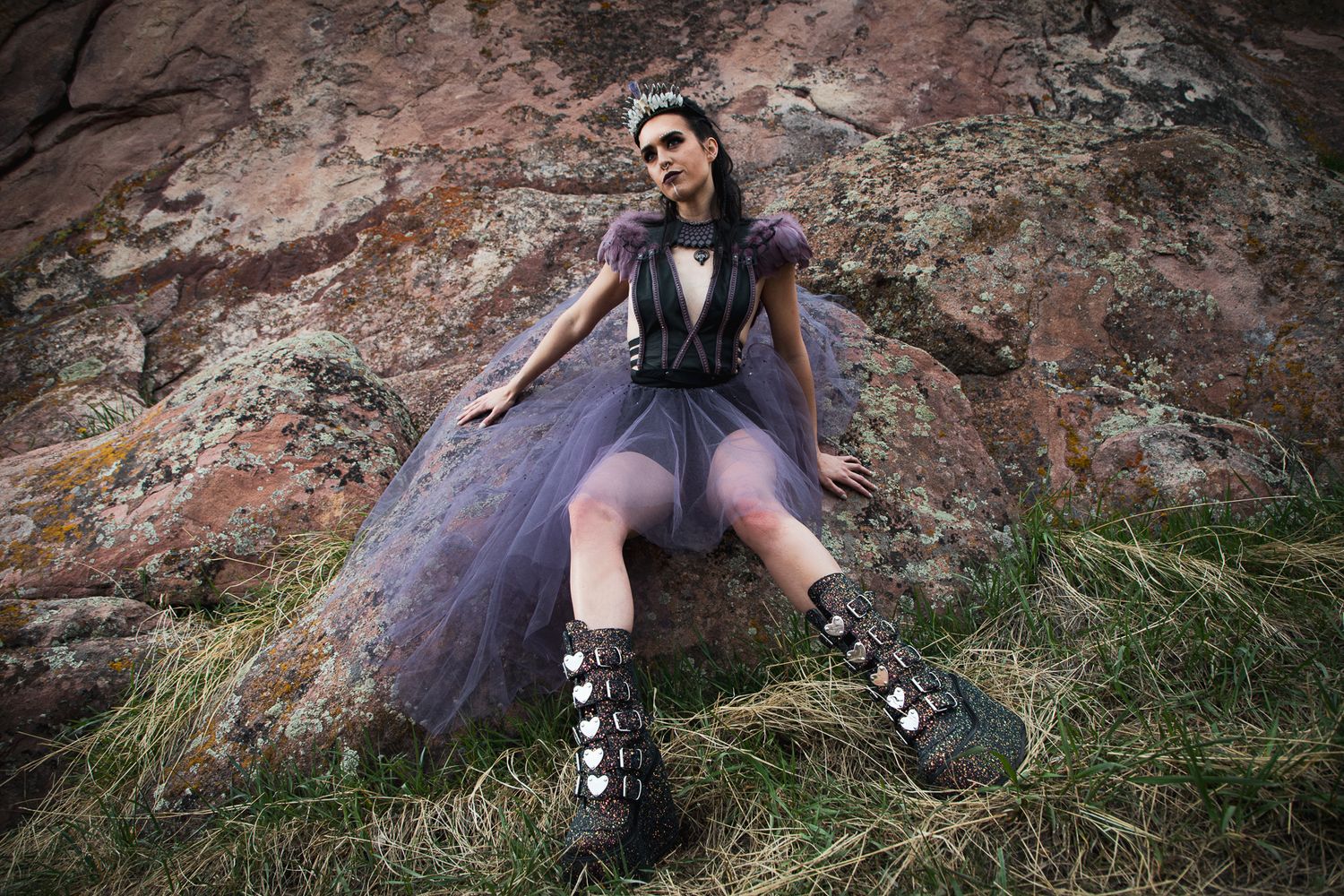 Gothic fashion model in purple tulle skirt and corset poses dramatically against rocky backdrop.