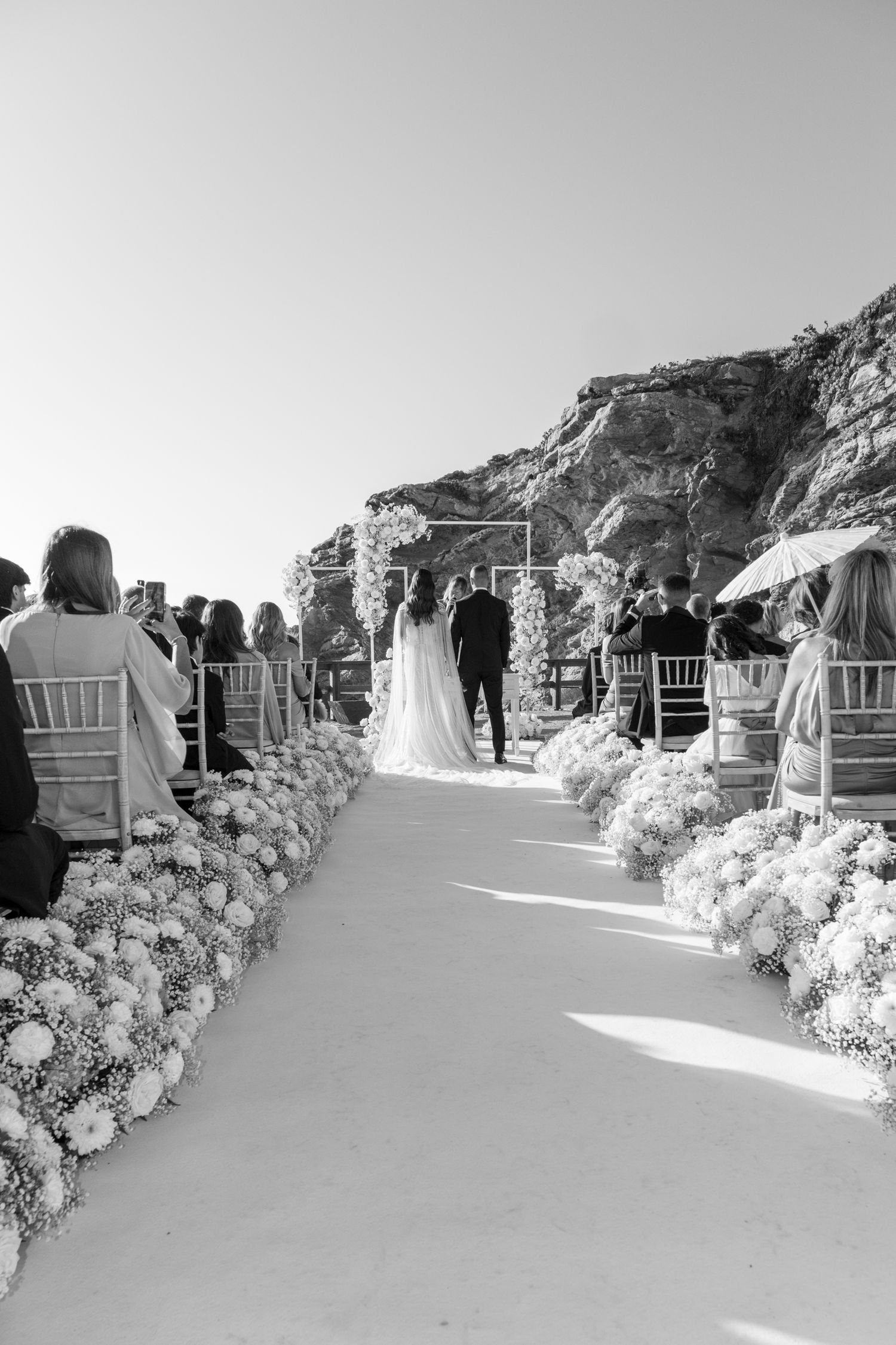 An outdoor wedding ceremony setup with white chairs and floral-lined aisle against dramatic cliffs.