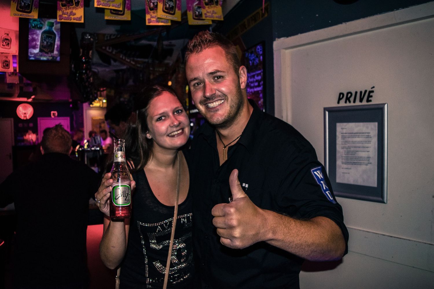 Two people posing together at a dimly lit bar with colorful posters on the wall.