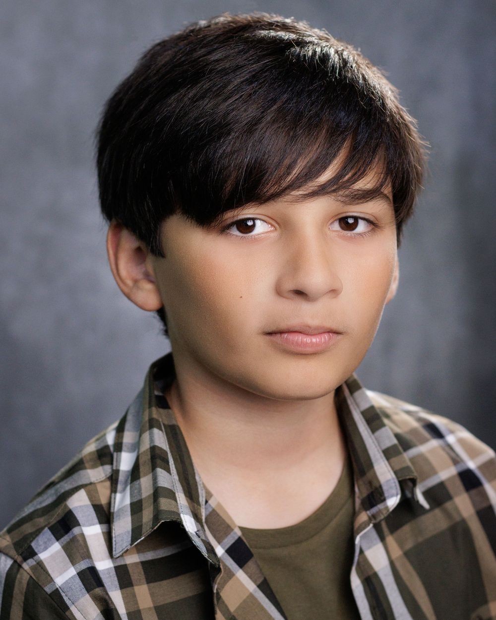 Portrait of a young person in a plaid button-up shirt over a dark t-shirt against a grey studio backdrop.