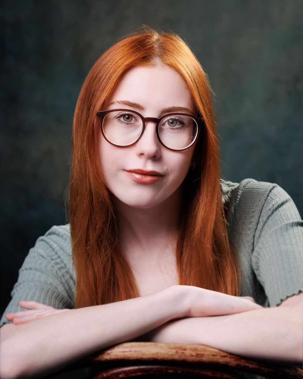 A person with long red hair and glasses rests their head on folded arms against a dark background.