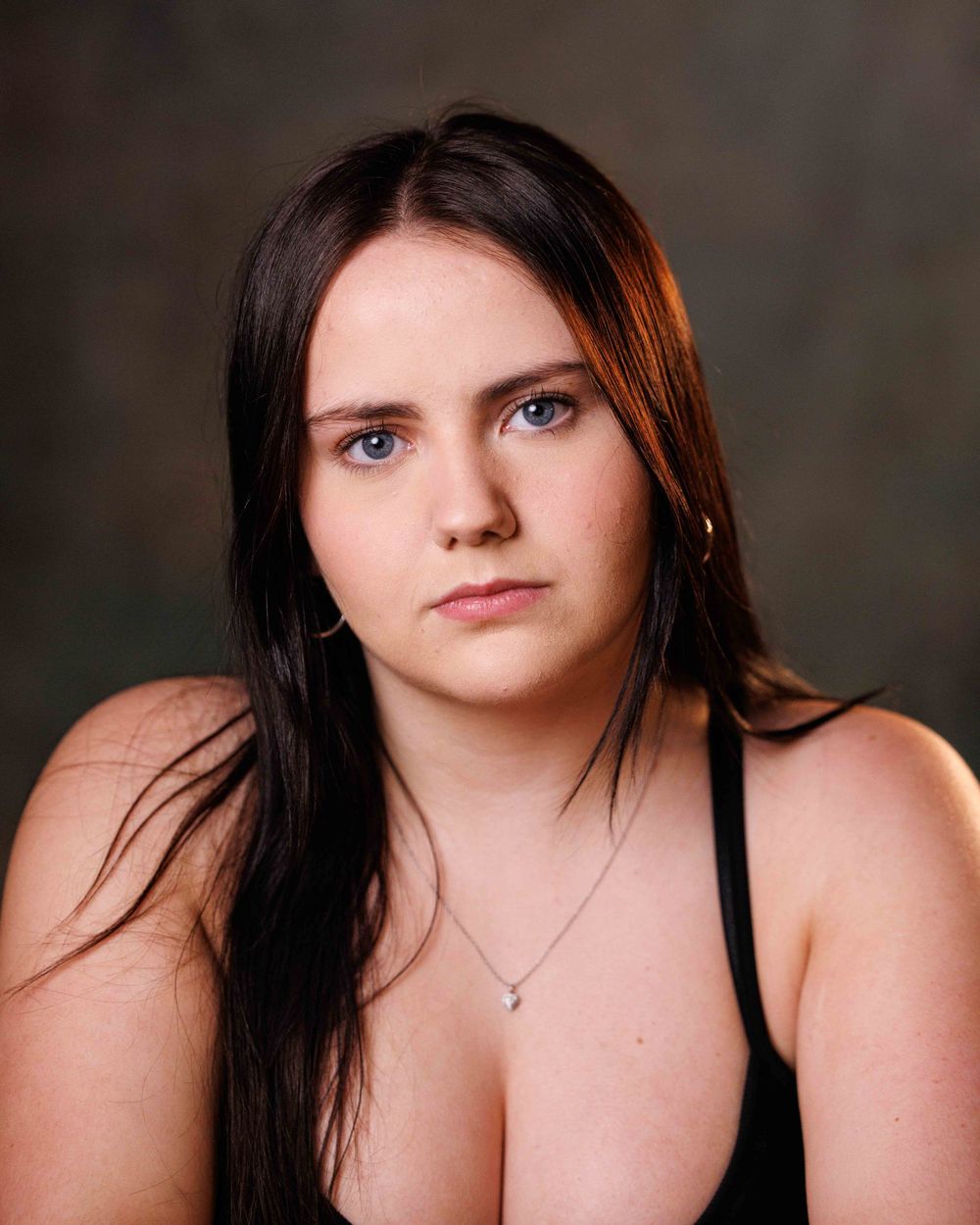 Young woman with long dark hair and blue eyes wearing a black top and silver necklace poses for a portrait photo.