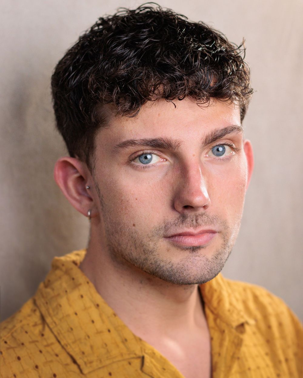 A person wearing a mustard yellow patterned shirt poses against a beige wall with bright blue eyes.