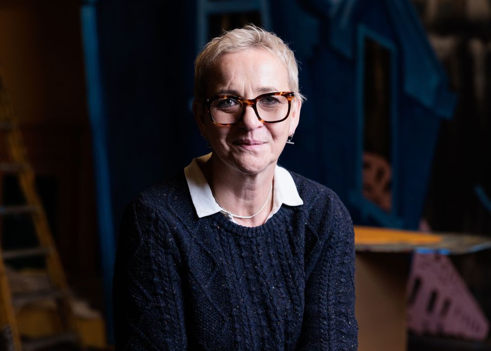 A person in glasses and a dark sweater poses against a blurred background in a professional headshot series.