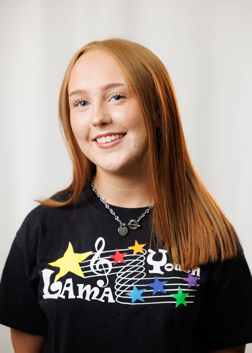 Smiling teenage girl with red hair wearing a black LAMIA Youth t-shirt with colorful stars and music notes.