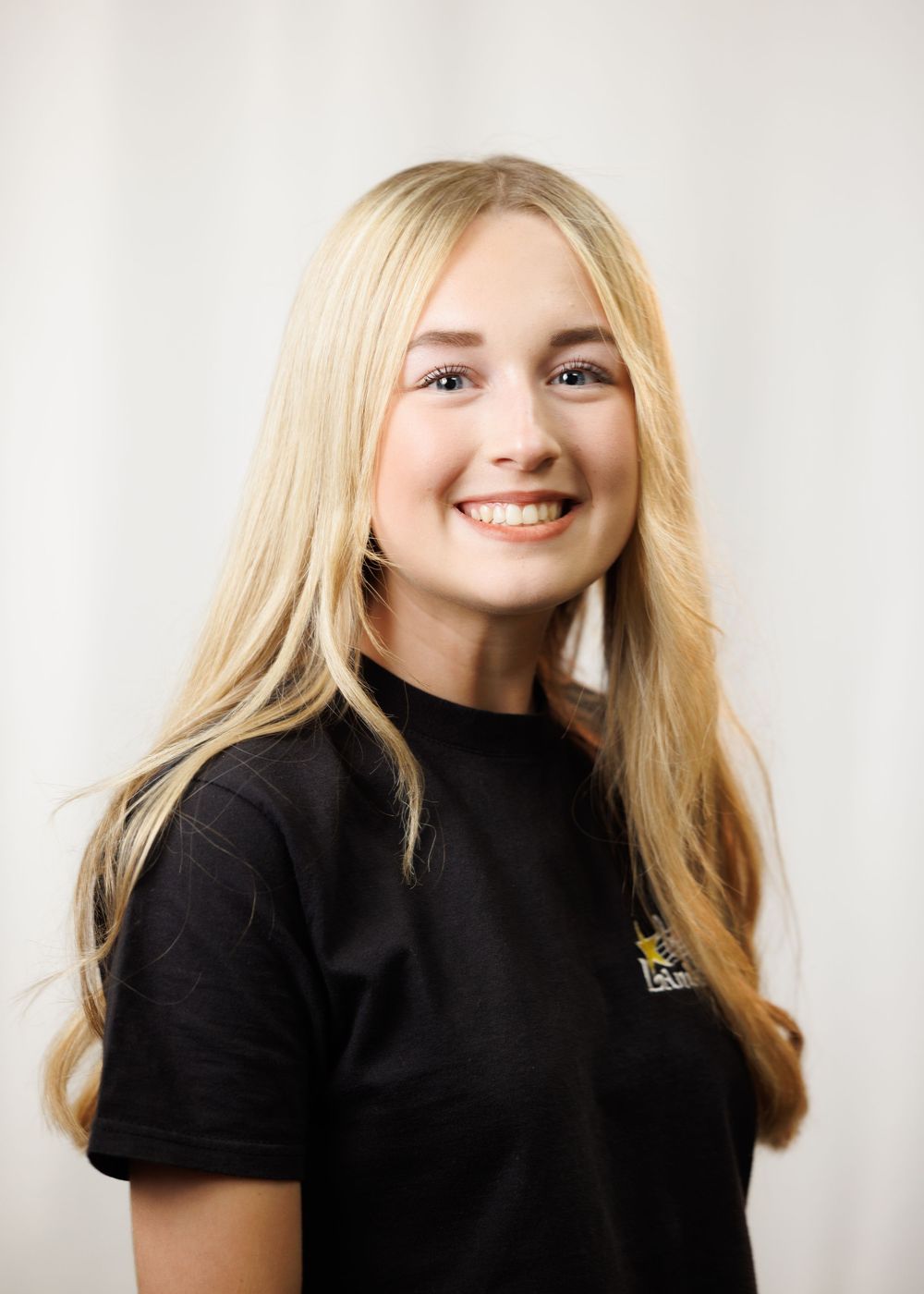 Young blonde woman smiling in black t-shirt with logo, professional headshot photo against white background.