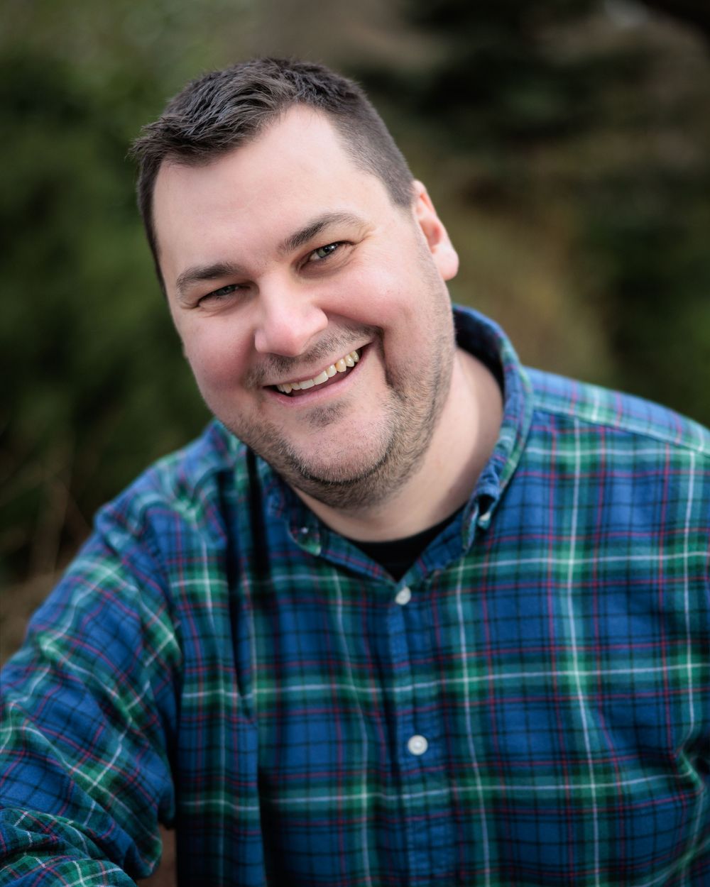 A person wearing a blue and green plaid button-down shirt smiles warmly at the camera in an outdoor setting.