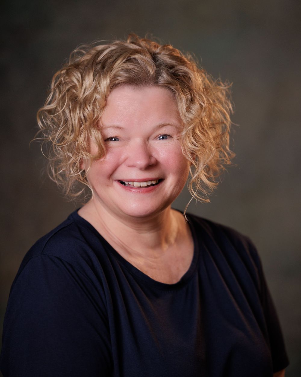 Professional headshot of a smiling woman with curly blonde hair wearing a navy top against a neutral background.