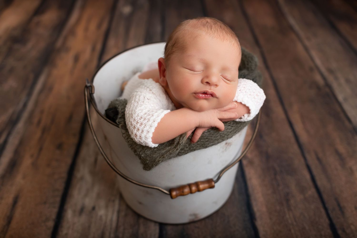 Sleeping newborn nestled in white bucket prop on wooden floor.