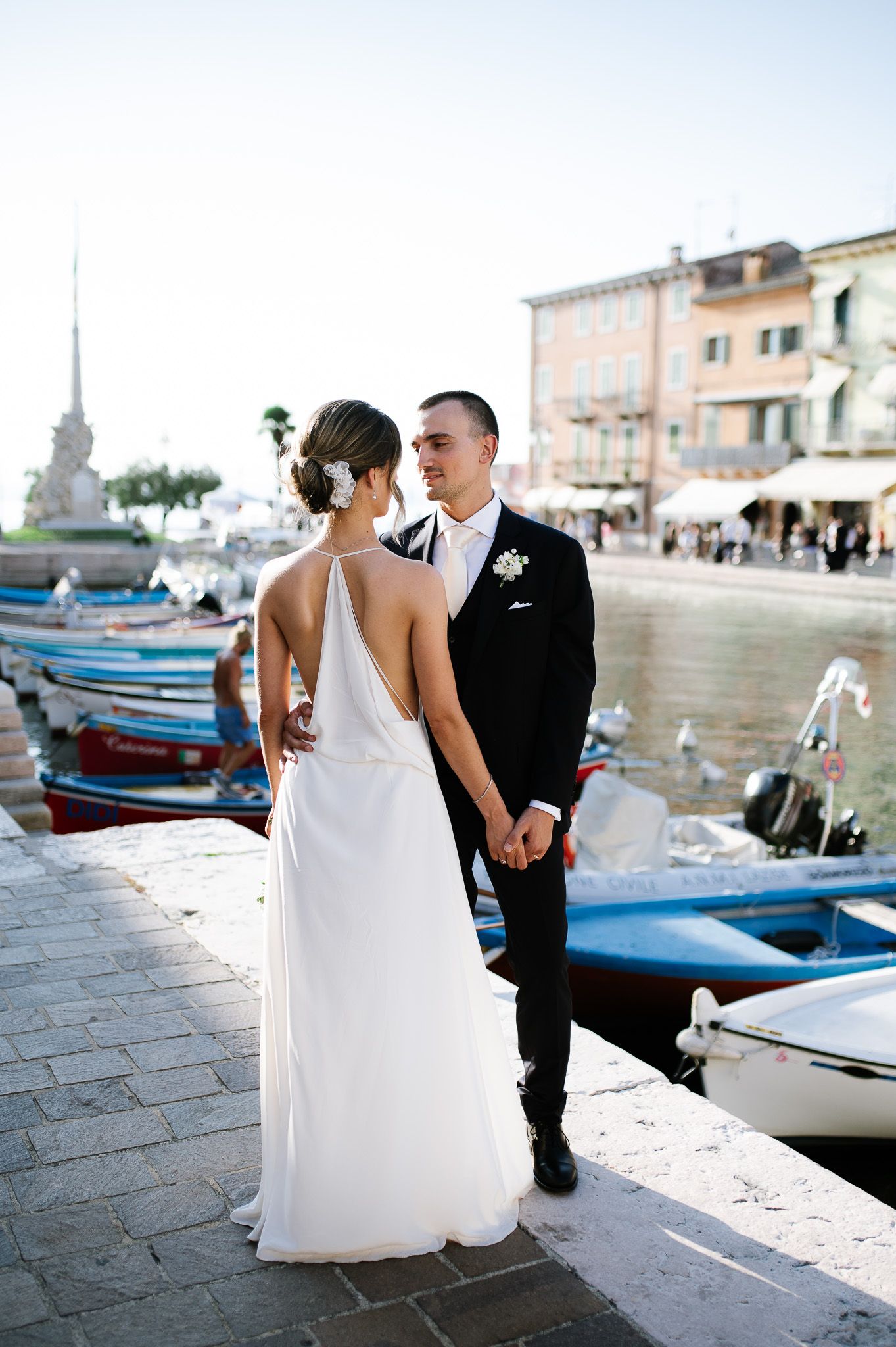 Bride and groom pose by a scenic Italian harbor with colorful boats, bride in elegant backless white gown.