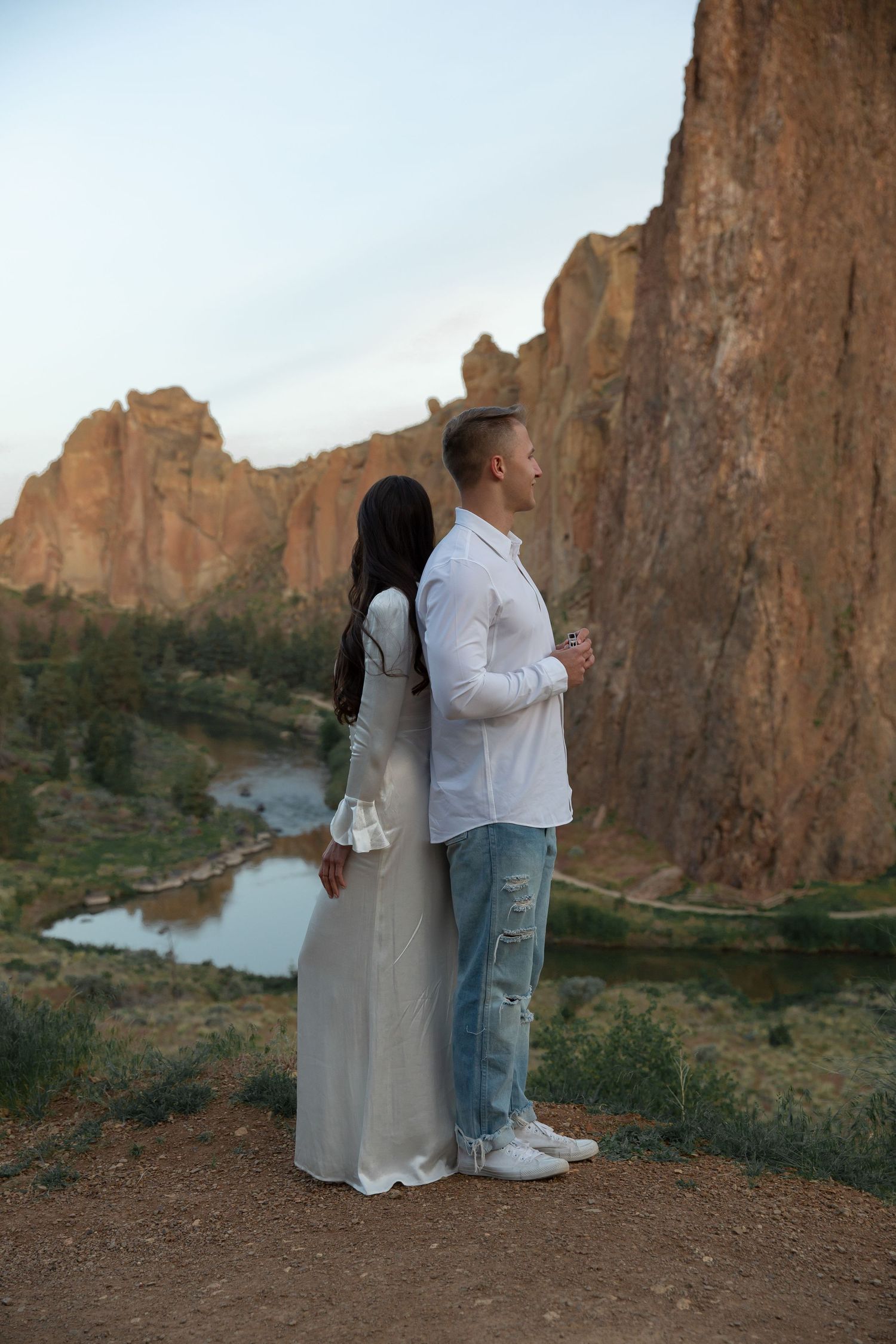 Couple stands together on cliff edge overlooking scenic river and rocky landscape at Smith Rock State Park in Oregon.