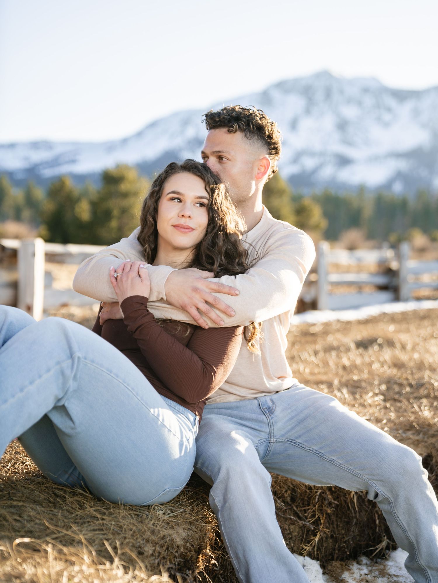 A couple sits together on hay bales with snow-capped mountains in the background during golden hour.