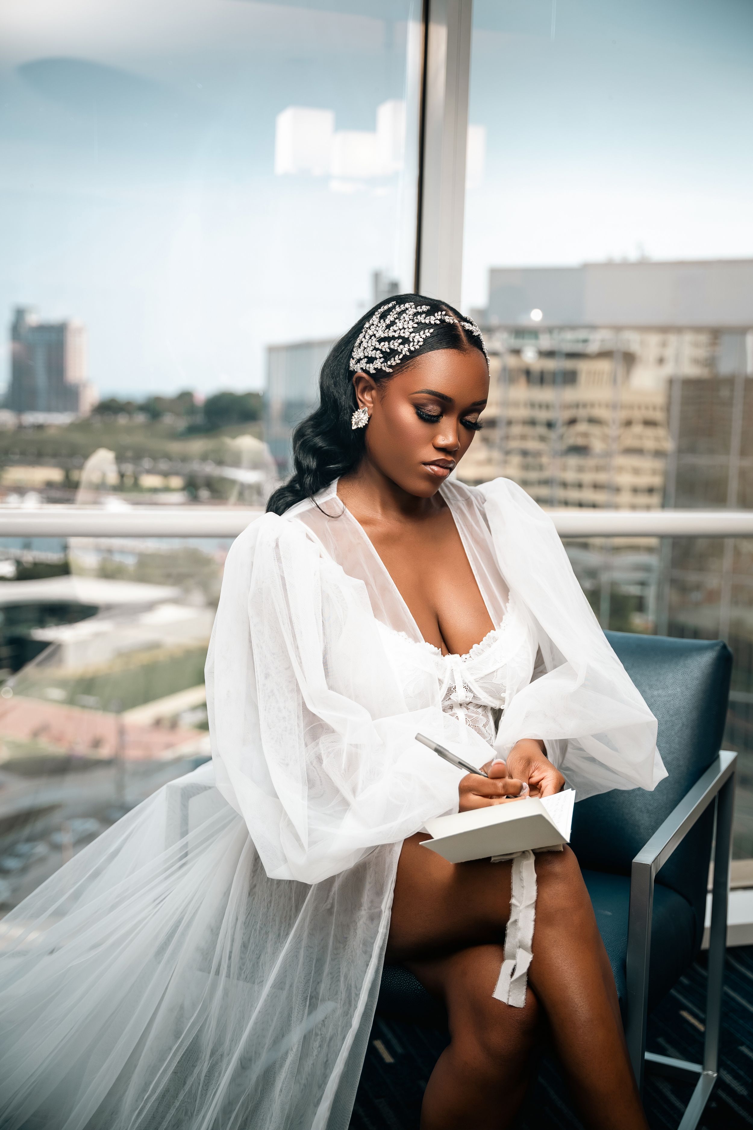 Bride in white robe and headwrap poses elegantly by window with city skyline view.