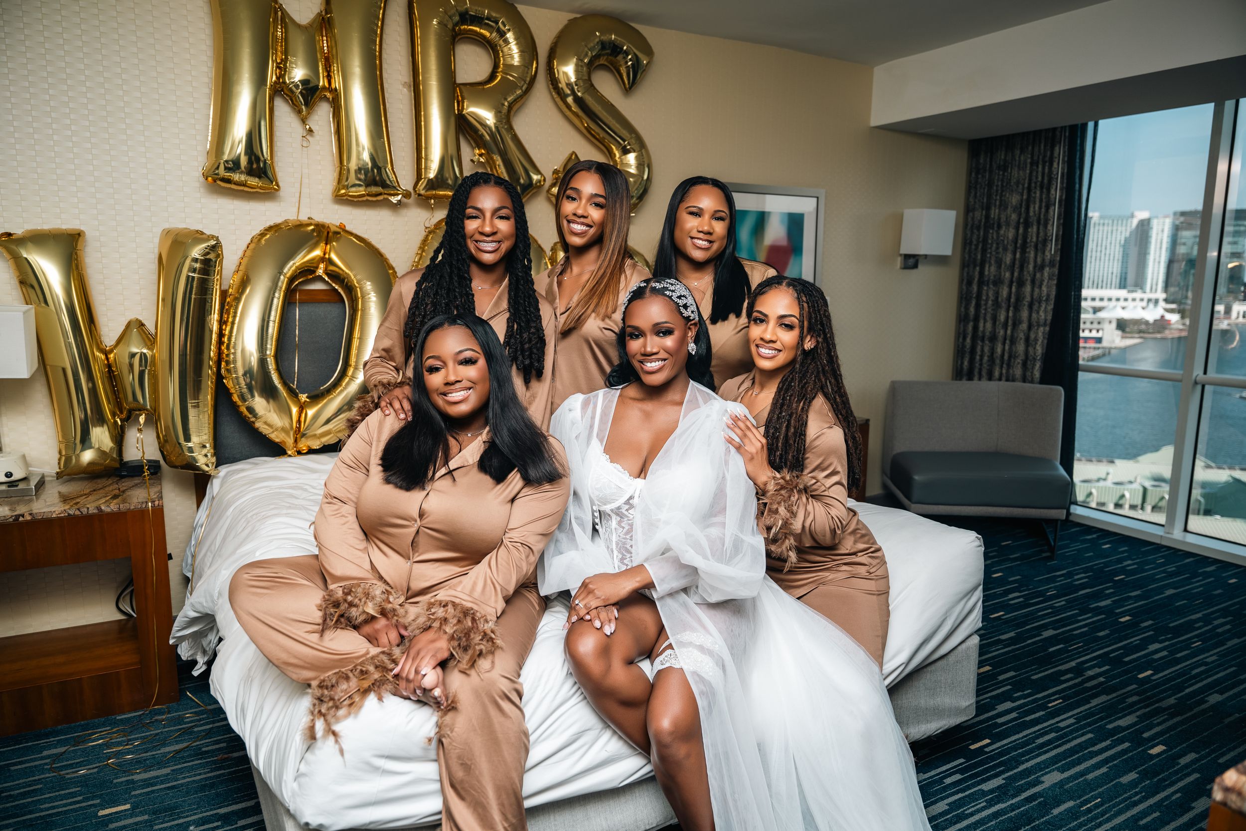 Wedding party in matching robes poses on hotel bed with gold MRS balloon decorations.