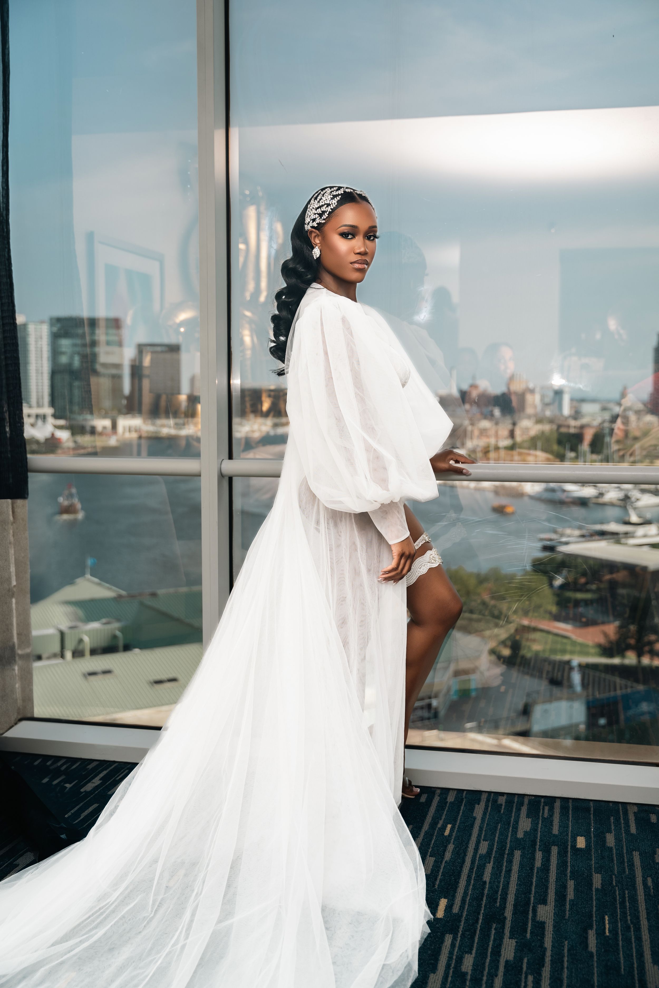 Bride in white robe poses by window overlooking harbor and city buildings.