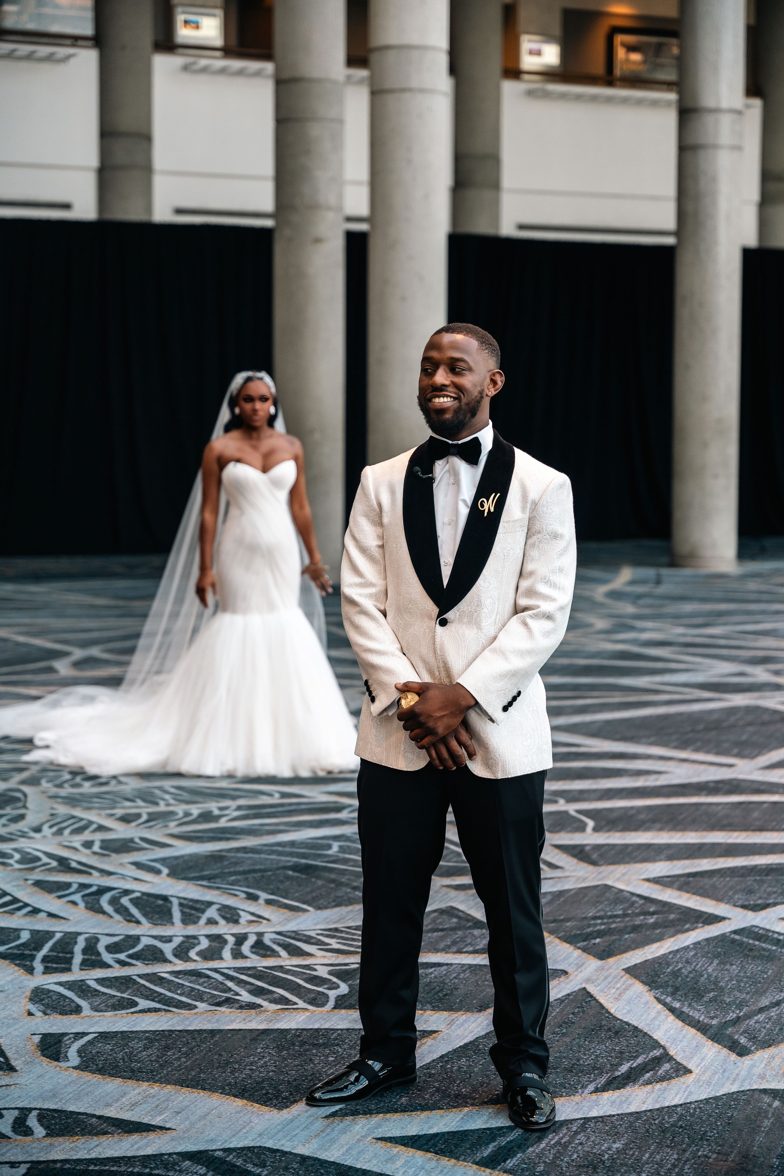 First look wedding photo with groom in white tuxedo and bride in background.