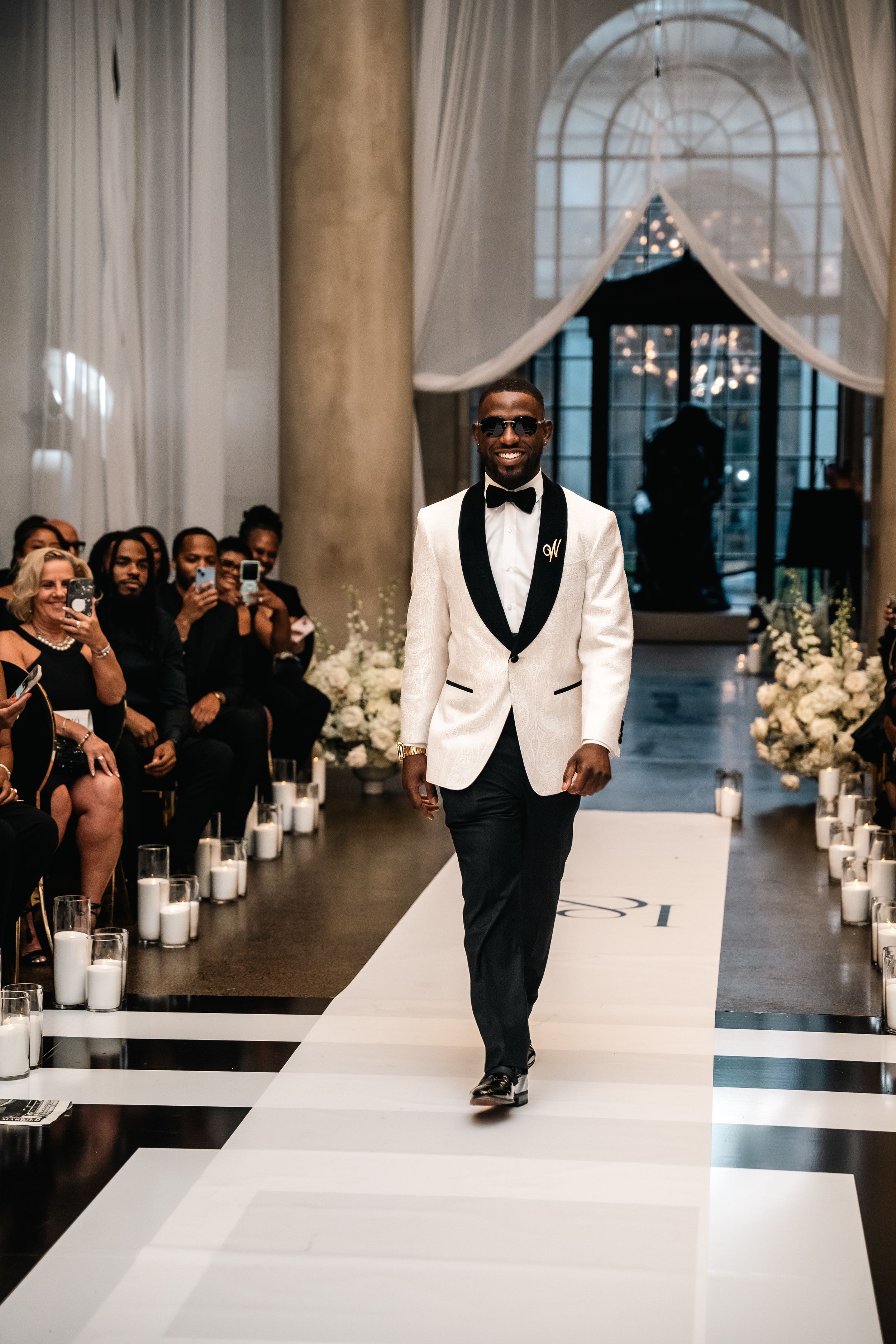 Groom walks down black and white aisle runner in white tuxedo jacket with candle decorations.