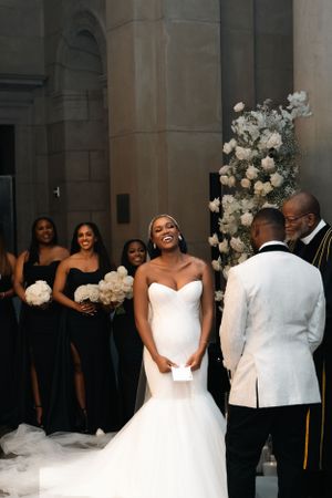 Wedding party stands in church as bride in white mermaid gown laughs joyfully during ceremony.