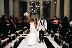 Wedding couple walks down aisle with white floral decor and string lights overhead.