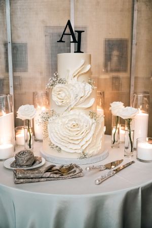 Elegant white wedding cake with large sugar roses and monogram topper surrounded by candles.