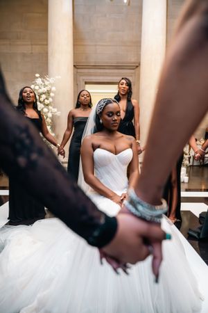 Bride in strapless gown sits while surrounded by bridesmaids in black dresses.