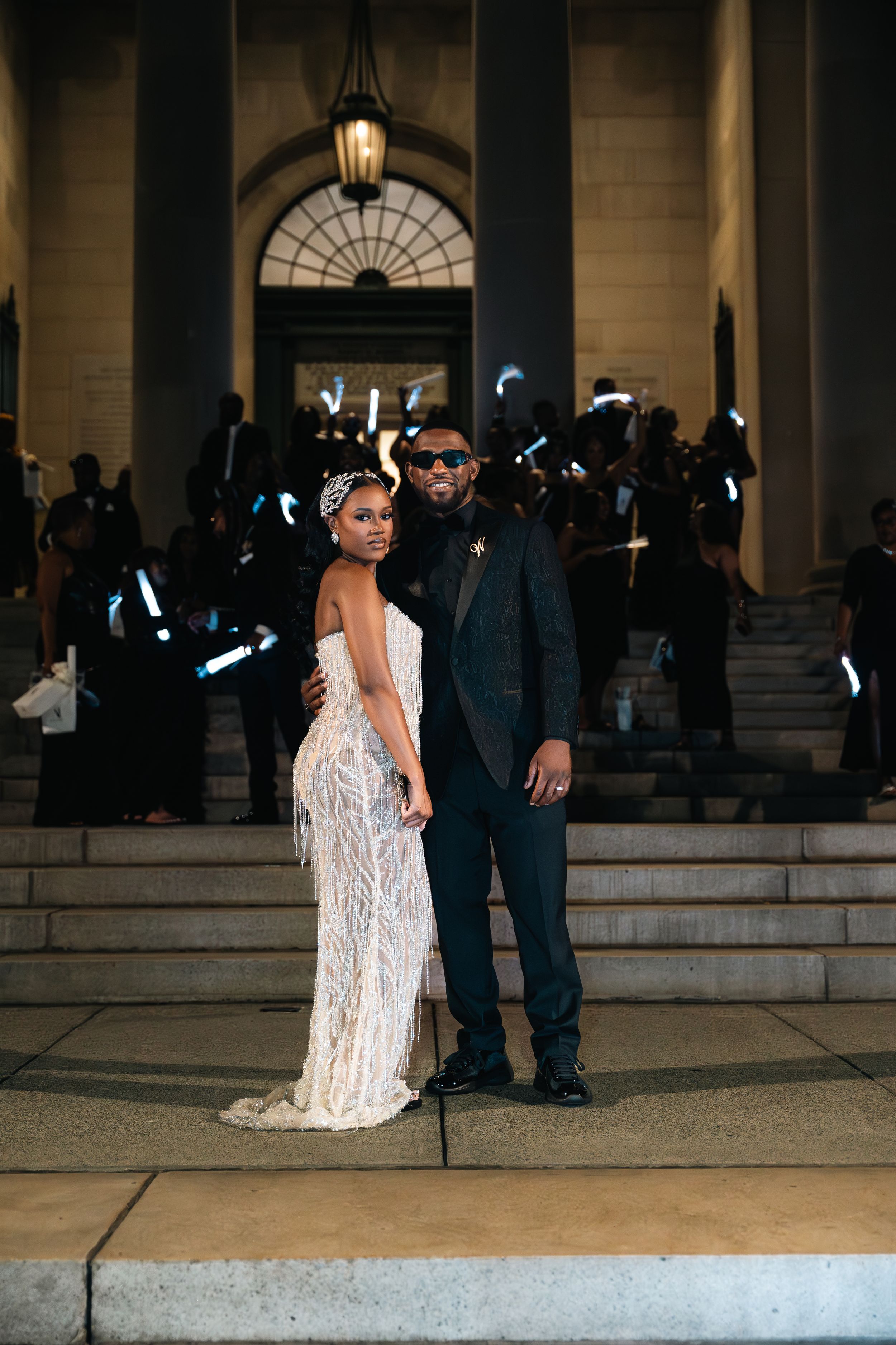 Elegant couple poses on grand stone steps at night with a beautifully lit archway behind them.