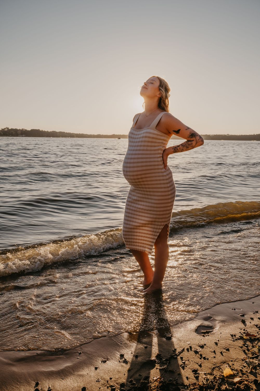 Pregnant woman in striped dress stands at water's edge during golden hour sunset at the beach.